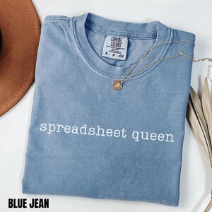 May include: A blue jean colored t-shirt with a crew neckline and the text spreadsheet queen printed in white across the chest. The shirt features a Comfort Colors brand label at the inner neck and is paired with a gold tone necklace with a small pendant.