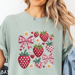 May include: A light green t-shirt featuring a strawberry design. The design includes red strawberries with white polka dots, pink bows, white daisies, and red hearts. The shirt has a relaxed fit.