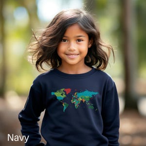 May include: A navy blue sweatshirt featuring a colorful world map design. The sweatshirt has a crew neck and long sleeves. The word "Navy" is printed at the bottom left corner of the image. The background is a blurred outdoor scene.