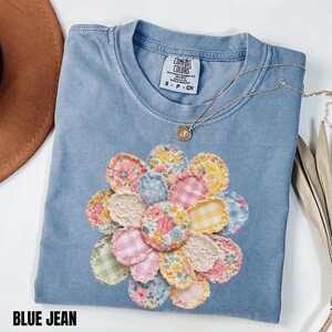 Könnte beinhalten: Ein blaues Jeans-T-Shirt mit einem floralen Patchwork-Design. Das Shirt hat einen Rundhalsausschnitt und kurze Ärmel. Der Text "BLUE JEAN" befindet sich am unteren Rand des Bildes. Eine goldene Halskette mit Anhänger befindet sich auf dem Shirt.