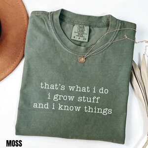 May include: A moss green t-shirt with the text "that's what I do, I grow stuff and I know things" in white. The shirt has a crew neck and short sleeves. A gold necklace with a pendant is draped on the shirt.