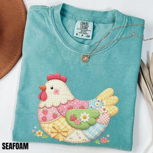 May include: Seafoam green t-shirt with a patchwork chicken design. The chicken features pink, yellow, and blue patterned sections, with strawberry and flower accents. A gold necklace with a pendant rests on the shirt. The shirt has a Comfort Colors tag.