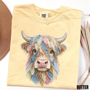 May include: Pale yellow t-shirt with a Highland cow design. The cow's fur is decorated with colourful patchwork. The t-shirt has a crew neck and the brand name "COMFORT COLORS" is visible on the tag. A delicate star pendant necklace rests on the shirt.
