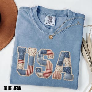 May include: A blue jean coloured t-shirt with the word "USA" appliquéd on the front. The letters are made of patchwork fabric in red, white, and blue patterns. A gold necklace with a pendant is draped on the shirt. The text "BLUE JEAN" is at the bottom.