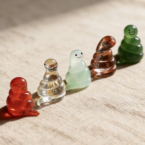 Puede incluir: Una fila de cinco figuritas de cristal de colores con forma de pequeños gusanos estilizados. Las figuritas son de color rojo, transparente, verde claro, marrón y verde oscuro. Cada una tiene una cara sonriente simple.