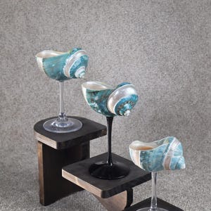 May include: Three unique wine glasses with seashell bowls, each perched on a small, dark wooden stand. The shells are a mix of teal, white, and silver, creating a coastal theme. The stems are clear and black.