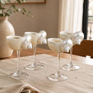 May include: A set of four stemmed glasses, each featuring a bowl crafted from an iridescent, pearlescent seashell. The shells are attached to clear glass stems and circular bases, creating a unique vessel design with a smooth, glossy finish.