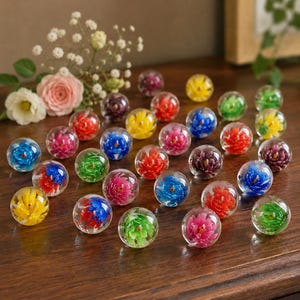 Puede incluir: Una colección de esferas de vidrio transparente, cada una con un diseño floral vibrante y multicolor. Las esferas están dispuestas en un patrón disperso, con varios colores que incluyen pétalos rojos, azules, amarillos, verdes y morados encapsulados dentro del vidrio.