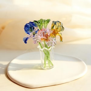 May include: A glass floral arrangement featuring three colourful iris flowers in a small glass vase. The irises are blue, pink, and yellow, with green stems and leaves. The vase sits on a white, oval-shaped ceramic plate.