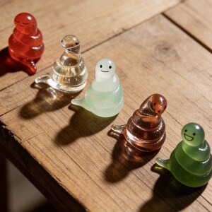 Może przedstawiać: Pięć kolorowych szklanych figurek ślimaków z uśmiechniętymi twarzami ułożonych na drewnianej powierzchni. Ślimaki są w odcieniach czerwieni, przezroczystości, jasnozielonego, bursztynowego i ciemnozielonego. Każdy ślimak ma spiralny korpus i mały ogon.