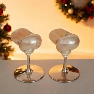 Puede incluir: Dos elegantes copas de vino con un diseño único. Los cuencos tienen forma de conchas iridiscentes, sostenidas por finos tallos y bases redondas de plata pulida. El fondo es suave y cálido, lo que sugiere una ocasión festiva o celebración.