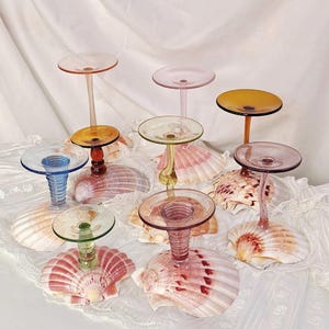 May include: A collection of eight glass candle holders, each featuring a unique colored glass stem attached to a natural pink and white scallop seashell base. The stems vary in color, including blue, green, amber, pink, and clear glass.