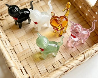 May include: Five small glass cat figurines in black, white, amber, pink, and light green. Each cat has a smooth, glossy finish, a pointed tail, and a small red accent at the neck. The figurines are arranged inside a woven light brown wicker basket.