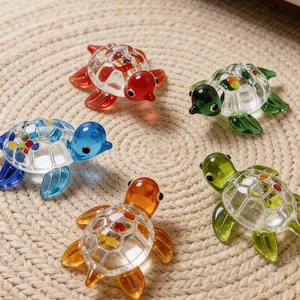 May include: Five glass turtle ornaments in various colours. Each turtle has a clear body with coloured details and coloured heads and flippers. They are displayed on a beige woven mat.