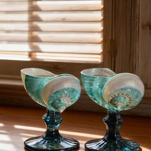 May include: Two decorative teal and white seashell-shaped goblets with dark teal stems and bases. The goblets are made of glass and sit on a wooden surface, with a window and blinds in the background.