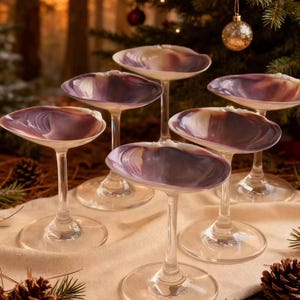 Op de afbeelding: Zes wijnglazen met een uniek ontwerp. De kommen zijn schelpvormig met een paarse en witte kleurovergang. De stelen en voetjes zijn van helder glas. De glazen staan op een crèmekleurige doek, met dennenappels en groen in de voorgrond.