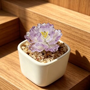May include: A small, square, white ceramic pot filled with small, light-coloured pebbles. A delicate, glass flower with purple petals and a yellow centre sits atop the pebbles. The flower is the focal point, on a wooden surface.