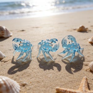 Puede incluir: Tres figuras de cangrejos azules transparentes con lunares blancos están dispuestas en una playa arenosa, con conchas marinas y una estrella de mar. Los cangrejos tienen pinzas y patas detalladas, y el fondo muestra el océano bajo un sol brillante.