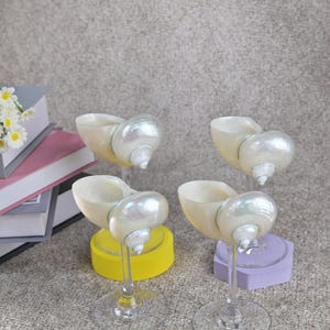 May include: Four decorative wine glasses with seashell-shaped bowls and pearl-like accents. The glasses have clear stems and bases, and are set on yellow and purple geometric bases. The seashells are off-white with iridescent highlights.
