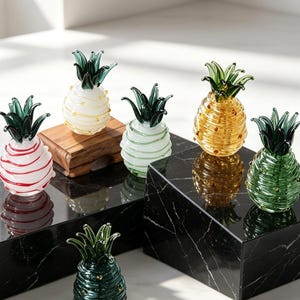 May include: A collection of decorative glass pineapple figurines in various colors, including white, red, green, and gold. Each pineapple has a green leafy top and is displayed on a black marble surface. The figurines are approximately 4 inches tall.