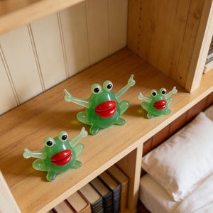 May include: Three green frog toys with red lips and outstretched arms are displayed on a wooden shelf. The frogs are of varying sizes, with the largest in the center. The shelf is part of a bookcase, with books visible below.