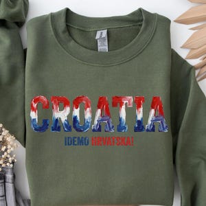 A green sweatshirt with the word "Croatia" printed on it, along with the text "IDEKO HVRVATSKA!" printed below. The sweatshirt is displayed on a white background, with some decorative leaves and a green fabric item visible in the background.