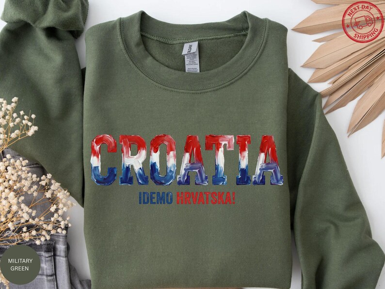 A green sweatshirt with the word "Croatia" printed on it, along with the text "IDEKO HVRVATSKA!" printed below. The sweatshirt is displayed on a white background, with some decorative leaves and a green fabric item visible in the background.