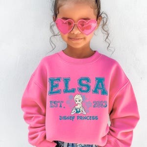 A young girl wearing a pink sweater with the text "ELSA EST. 2013 DISNEY PRINCESS" and heart-shaped sunglasses.