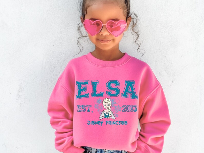 A young girl wearing a pink sweater with the text "ELSA EST. 2013 DISNEY PRINCESS" and heart-shaped sunglasses.