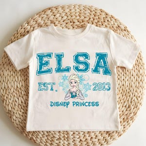 A white t-shirt with a design featuring the character Elsa from the Disney movie Frozen, along with the text "ELSA EST. 2013 DISNEY PRINCESS".