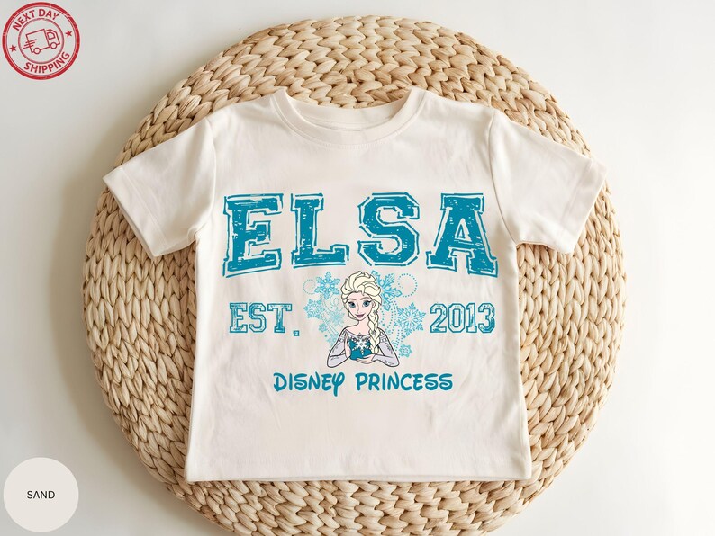 A white t-shirt with a design featuring the character Elsa from the Disney movie Frozen, along with the text "ELSA EST. 2013 DISNEY PRINCESS".