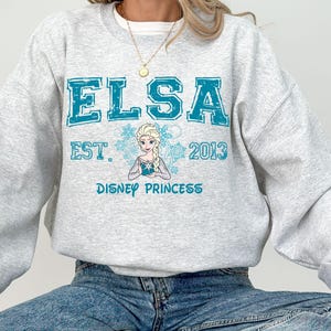 A person wearing a gray sweatshirt with a design featuring the character Elsa from the Disney movie Frozen.