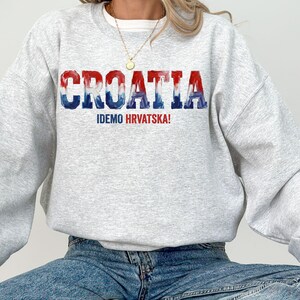 A person wearing a grey sweatshirt with the word "Croatia" printed on it, along with the text "IDEMO HRVATSKA!" printed below.