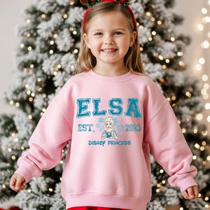 A young girl wearing a pink sweatshirt with the text "ELSA EST. 2013 DISNEY PRINCESS" is standing in front of a Christmas tree.