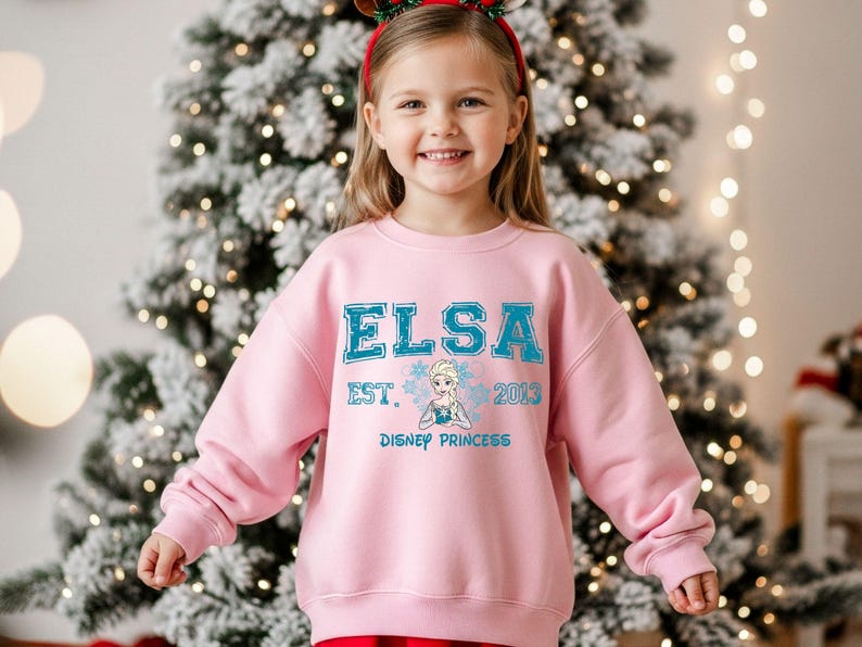 A young girl wearing a pink sweatshirt with the text "ELSA EST. 2013 DISNEY PRINCESS" is standing in front of a Christmas tree.