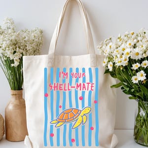 May include: Cream-colored tote bag with a cartoon turtle design. The design features a yellow turtle with an orange shell, blue vertical stripes, and the text "I'M YOUR SHELL-MATE" in pink and red. The bag has two handles.