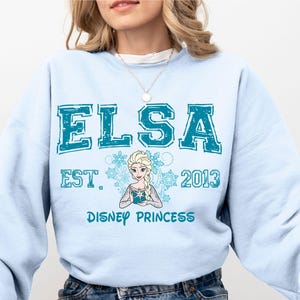 A woman wearing a light blue sweatshirt with the text "ELSA EST. 2013 DISNEY PRINCESS" printed on it.