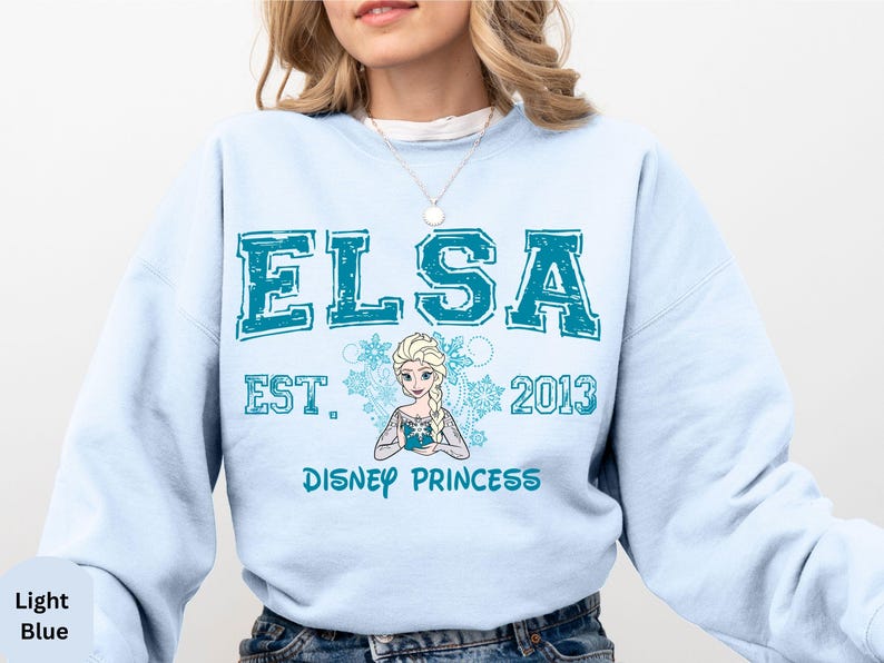 A woman wearing a light blue sweatshirt with the text "ELSA EST. 2013 DISNEY PRINCESS" printed on it.