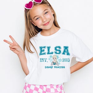A young girl wearing a white t-shirt with a graphic of Elsa from Frozen and the text "ELSA EST. 2013 DISNEY PRINCESS" is smiling and making a peace sign gesture.