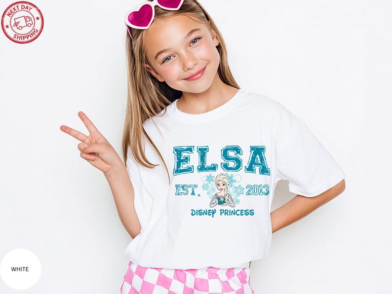 A young girl wearing a white t-shirt with a graphic of Elsa from Frozen and the text "ELSA EST. 2013 DISNEY PRINCESS" is smiling and making a peace sign gesture.