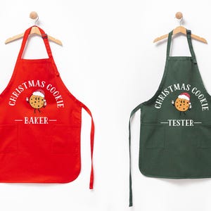 Puede incluir: Dos delantales, uno rojo y otro verde oscuro, colgando de perchas de madera. El delantal rojo dice "Christmas Cookie Baker" con un gráfico de galleta. El verde dice "Christmas Cookie Tester". Ambos tienen bolsillos.