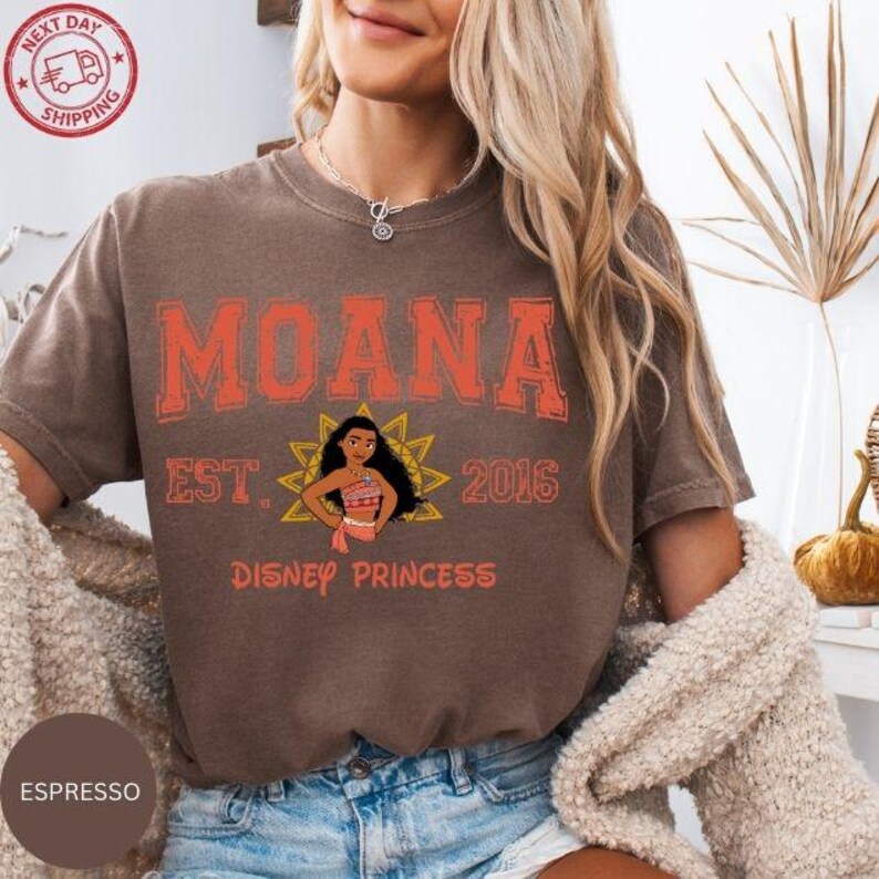 A woman wearing a brown t-shirt with the text "Moana Est. 2016 Disney Princess" printed on it, along with a graphic of Moana. She is also wearing a necklace and a beige sweater.