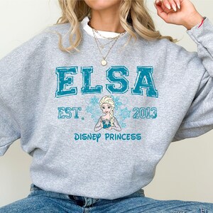 A person wearing a gray sweatshirt with the text "ELSA EST. 2013 DISNEY PRINCESS" printed on it.