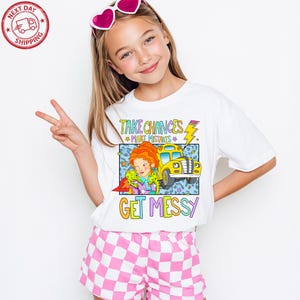 Puede incluir: Camiseta blanca con un gráfico colorido que presenta un autobús escolar amarillo y el texto "TAKE CHANCES, MAKE MISTAKES, GET MESSY". La camiseta se combina con pantalones cortos a cuadros rosas y blancos. Un logotipo de envío rojo y blanco está en la esquina superior izquierda.