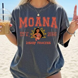 A person wearing a gray t-shirt with the text "Moana" and "Disney Princess" printed on it, along with a Disney princess graphic. The person is holding a drink in their hand.
