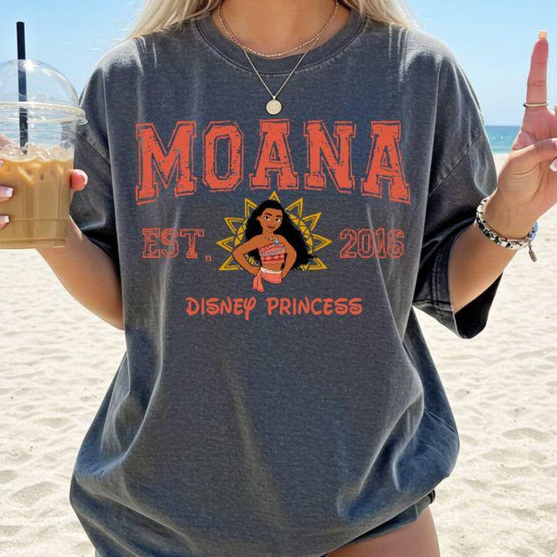A person wearing a gray t-shirt with the text "Moana" and "Disney Princess" printed on it, along with a Disney princess graphic. The person is holding a drink in their hand.