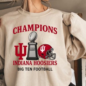 Puede incluir: Sudadera color arena con la palabra "CHAMPIONS" en rojo sobre un trofeo de fútbol americano y un casco rojo. Las palabras "INDIANA HOOSIERS" y "BIG TEN FOOTBALL" también están impresas en la sudadera.