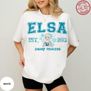A woman wearing a white t-shirt with the text "ELSA EST. 2013 DISNEY PRINCESS" printed on it, along with a depiction of the Disney princess Elsa.