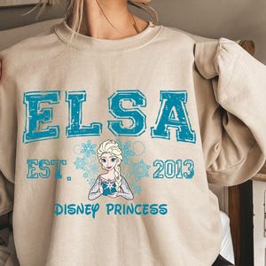 A person wearing a beige sweater with a graphic design featuring the character Elsa from the Disney movie Frozen.