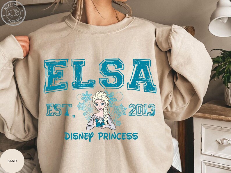 A person wearing a beige sweater with a graphic design featuring the character Elsa from the Disney movie Frozen.